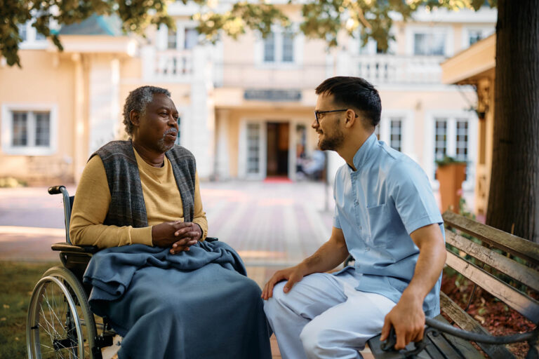 Solentra Wellness | Black senior in wheelchair talking to male nurse while relaxing in the park of residential care home.
